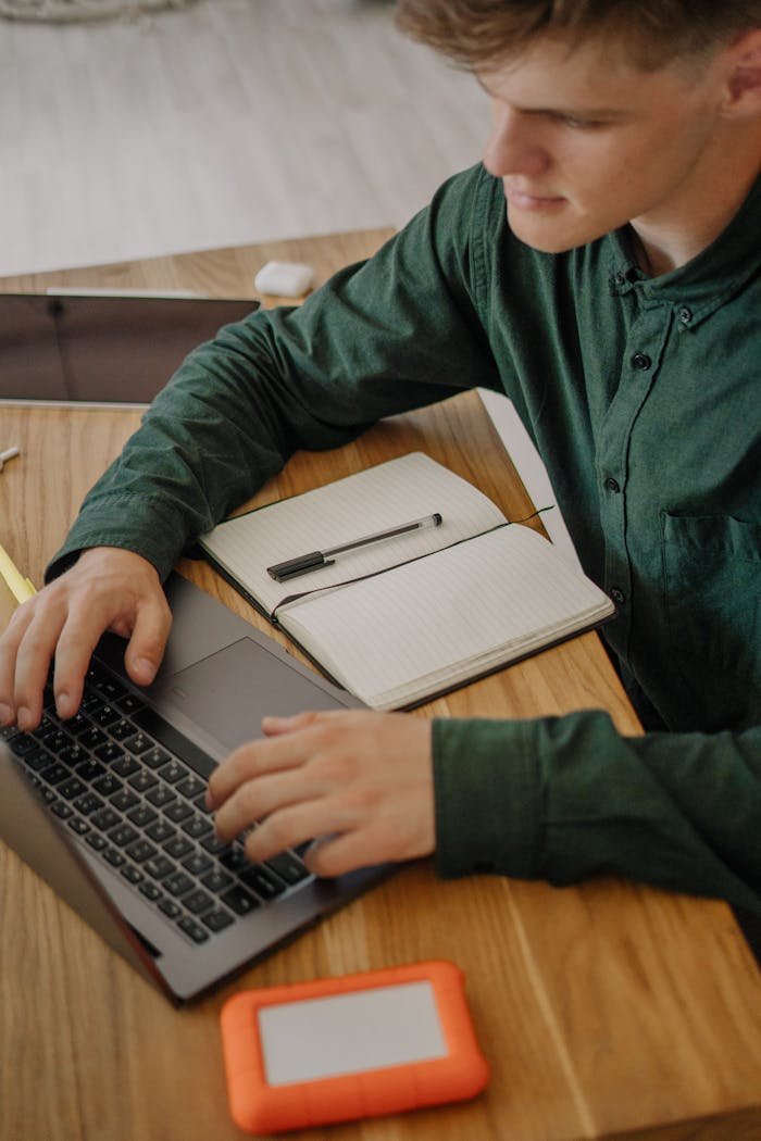 Young adult working on a laptop with notes on a wooden desk, focusing on online study or remote work.