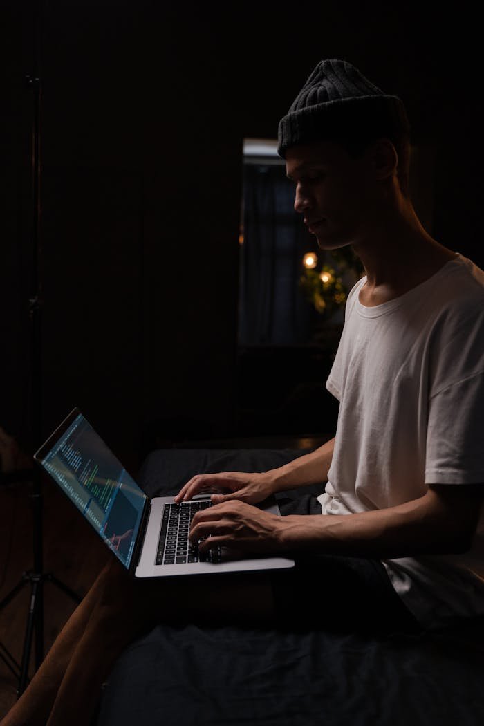 Side view of a young man in a beanie working on a laptop in a dark room, symbolizing remote work.
